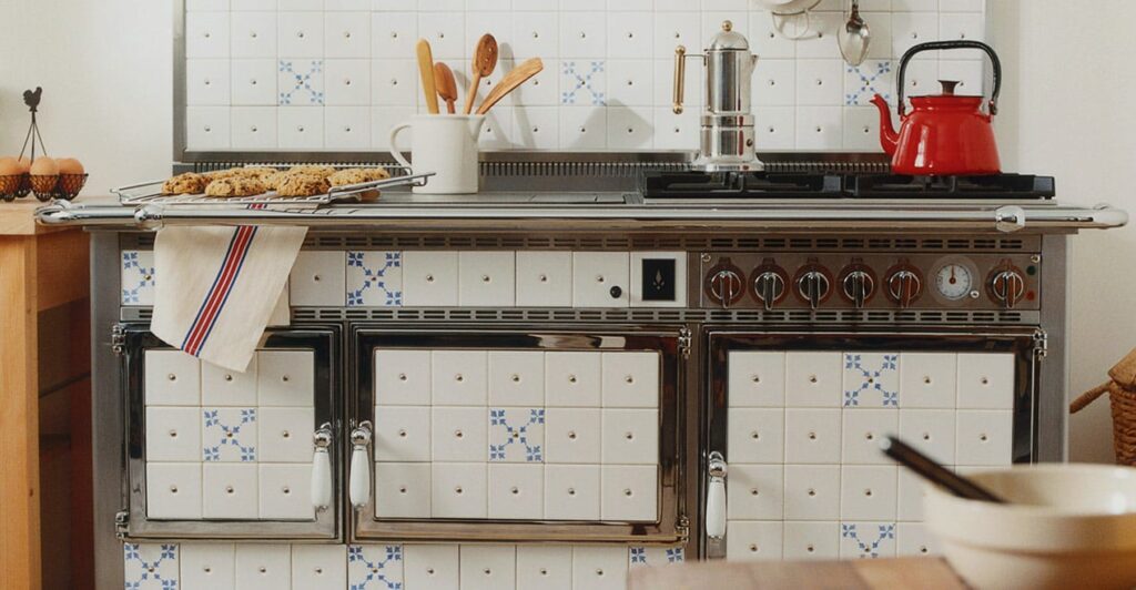 Une cuisine accueillante avec une cuisinière de style vintage avec des carreaux à motifs blancs et bleus. Sur la cuisinière se trouvent une bouilloire rouge et une cafetière en métal. Le plan de travail contient une tasse, divers ustensiles de cuisine et un plateau de biscuits. Un torchon blanc à motifs rayés est suspendu à la poignée du four.