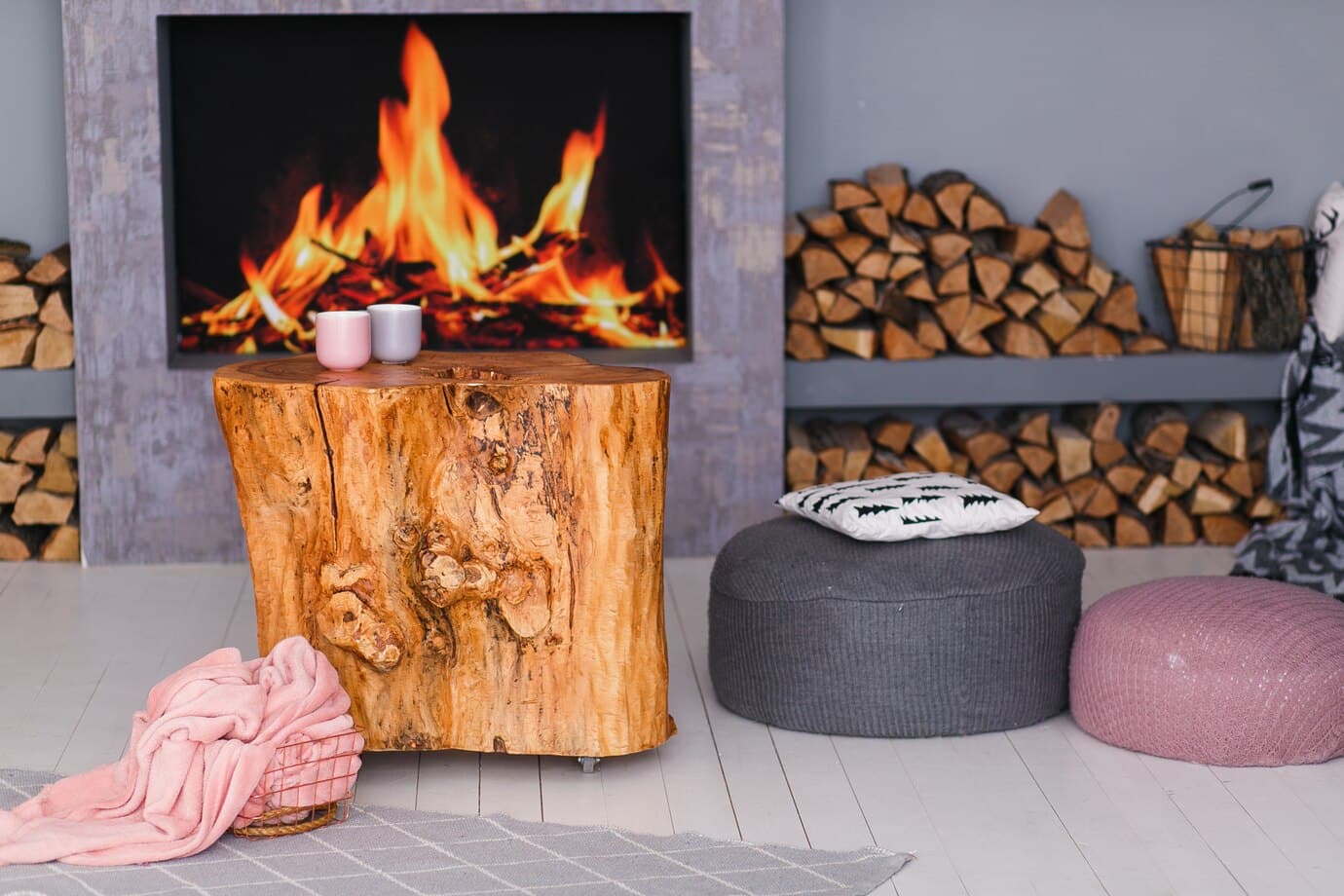 Un salon confortable est doté d'un poêle à bois allumé avec une pile de bois de chauffage de chaque côté. Devant la cheminée se trouve une table basse en souche de bois rustique. Une couverture rose, un pouf gris et un pouf rose entourent la table, avec un oreiller placé sur le pouf gris.