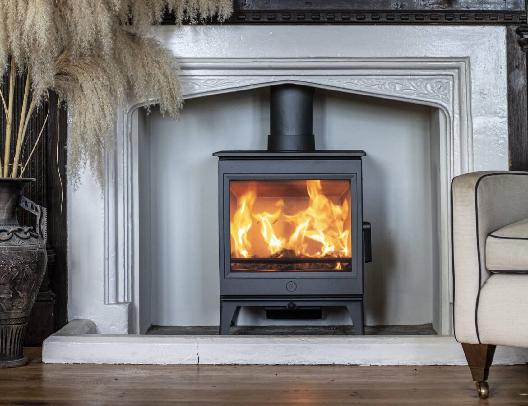 Un petit poêle à bois noir Charnwood, le Cranmore 7, avec un feu visible, est installé dans une cheminée blanche. Au-dessus de la cheminée se trouvent des panneaux en bois foncé détaillés ornés d'une composition florale. Un arrangement en pot d'herbe de pampa beige se trouve à gauche, et une partie d'une chaise beige est visible à droite.