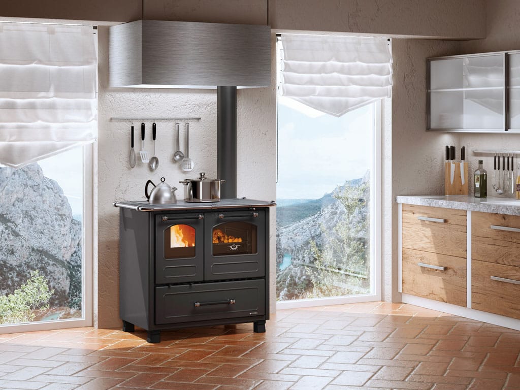 Une cuisine moderne comprend une cuisinière à bois NORDICA FAMILY 4.5 avec deux casseroles sur le dessus, située entre de grandes fenêtres avec vue sur la montagne. Des ustensiles sont suspendus au mur et des armoires en bois avec un comptoir en marbre se trouvent sur la droite. Des stores romains blancs couvrent les fenêtres, ajoutant une touche d'élégance.