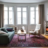 Un salon chaleureux avec un canapé vert, une table basse en bois et un fauteuil couleur crème sur un tapis à motifs rouges. Un poêle à bois Sergio Leoni allumé est placé à droite, à côté d'une table d'appoint. Trois grandes fenêtres avec des rideaux beiges illuminent l'espace.