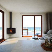 Un salon moderne avec un poêle à bois Rocal, un canapé et une table basse. De grandes portes vitrées s'ouvrent sur un patio donnant sur l'océan. La pièce présente un ANGLE de design minimaliste avec des murs et des sols de couleur claire. La lumière du soleil pénètre à travers les portes, renforçant la sensation d'espace.