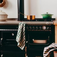 Une scène de cuisine chaleureuse avec une cuisinière Legende noire de style vintage avec une marmite blanche, une petite marmite en cuivre et une marmite verte sur les brûleurs. Une serviette à carreaux est drapée sur la porte du four. Au premier plan, un verre à vin et un torchon sont posés sur le comptoir. Des étagères avec des plats et une bougie sont à l'arrière-plan.