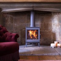 Une pièce chaleureuse est dotée d'un poêle à bois Charnwood Cranmore 5 allumé à l'intérieur d'une cheminée en pierre sous un manteau en bois. À gauche, une chaise rembourrée marron est partiellement visible. Trois bougies allumées sont placées sur le sol près de la cheminée. Un tapis à motifs recouvre le plancher en bois.