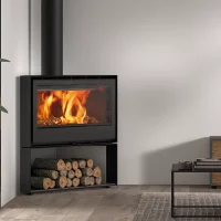 Un poêle à bois moderne, ROCAL HABIT 100 R, est installé dans le coin d'un salon, émettant une flamme orange vif. En dessous, un espace de rangement contient des bûches soigneusement empilées. À proximité se trouvent une porte vitrée, une plante en pot et une partie d'un canapé gris avec un coussin décoratif.