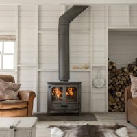 Un salon chaleureux avec un poêle à bois Charnwood Island III et un tuyau qui s'étend jusqu'au plafond. La pièce est dotée de murs en bois blanc, d'un tapis en peau de vache et de fauteuils en cuir. Le bois de chauffage est soigneusement empilé dans une alcôve à côté du poêle. Un coffre blanc fait office de table basse.