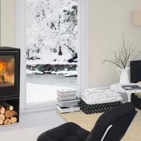 Un salon moderne présente un élégant poêle à bois ROCAL HABIT 50V avec un feu crépitant à l'intérieur. Les bûches sont stockées sous le poêle. La pièce comprend un canapé noir, une table d'appoint blanche, une pile de livres, un lampadaire et de grandes fenêtres affichant la scène extérieure enneigée sereine.