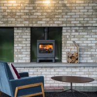 Un espace de vie confortable avec un poêle à bois Charnwood encastré dans un mur en briques. Un fauteuil bleu avec des coussins blancs et bordeaux se trouve à proximité, à côté d'une table basse ronde en bois. Il y a une petite pile de bois de chauffage à côté du poêle Skye 5, et deux grandes suspensions sont suspendues au-dessus.