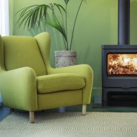 Un fauteuil vert rembourré est posé à côté d'un poêle à bois noir Charnwood Skye 7 avec un feu visible à l'intérieur. Le poêle est placé contre un mur vert clair avec une plante en pot en arrière-plan, et un tapis à motifs de couleur claire est posé sur le sol.