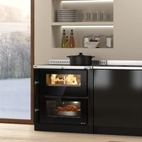 Une cuisine moderne est équipée d'une cuisinière Nordica Verona XXL, dont la surface noire épurée présente deux compartiments : celui du haut présente des flammes visibles suggérant qu'il s'agit d'une cuisinière à bois, et le compartiment inférieur contient un plat rôti. Au-dessus de la cuisinière se trouvent des étagères ouvertes ornées de plats et de bouteilles.