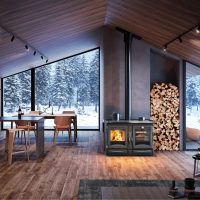 Un intérieur de cabine moderne avec de grandes fenêtres présentant une forêt enneigée, évoquant une sérénité digne de l'ALASKA. Il y a une table à manger avec des chaises, une cuisinière à bois avec un tas de bois de chauffage à côté, et un coin salon confortable avec des fauteuils et une table basse. L'espace présente des murs et des sols en bois.